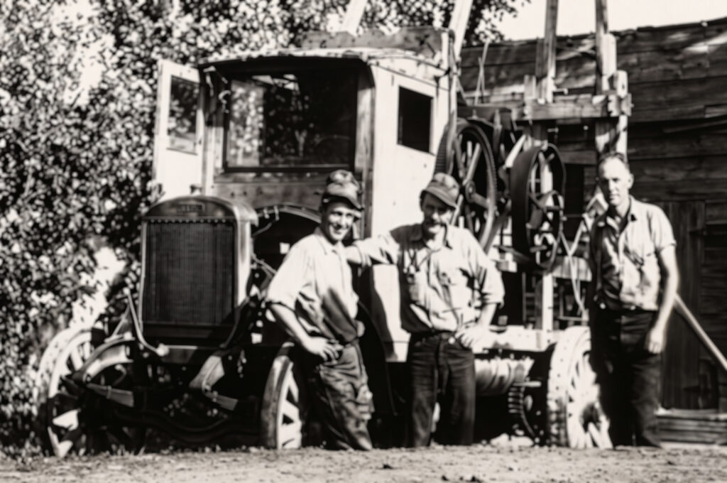 Three men stand in front of an old truck or machinery, posing and smiling for the camera. The scene is outdoors, with trees and a rustic wooden building in the background. The photo appears vintage or historical.