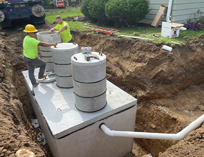 Two workers in safety gear install a large concrete septic tank with cylindrical risers in a dug-out area beside a house, with tools and pipes visible around the construction site.