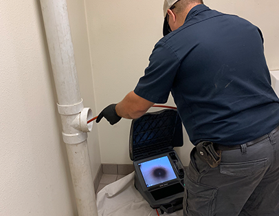 A person in work attire uses a pipe inspection camera to examine the inside of a white drainage pipe, with the camera monitor displaying a pipe interior.