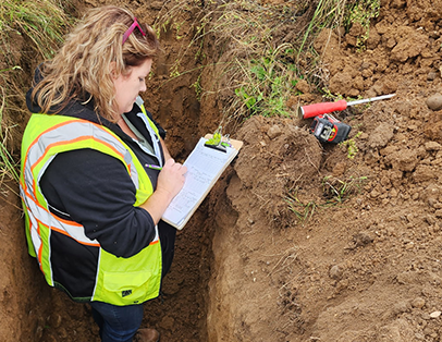 A woman wearing a reflective safety vest writes on a clipboard while standing in a soil trench with tools, including a soil auger and measuring tape, resting on the ground beside her.
