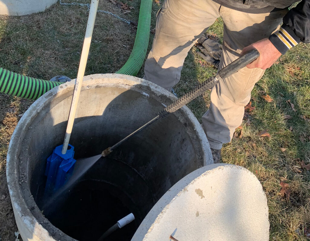 A person uses a pressure washer to clean the inside of a large concrete pipe or well outdoors. A green hose and a blue brush are also visible, along with grass and scattered leaves nearby.