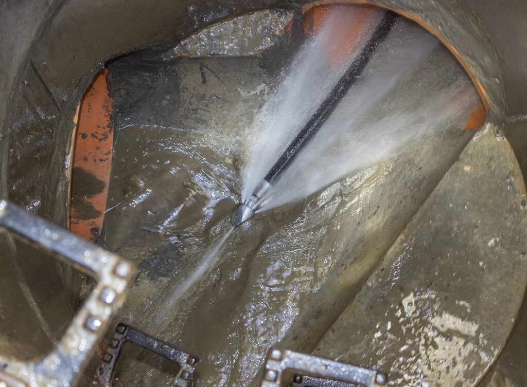 A high-pressure water jet cleans the inside of a large, muddy pipe or drain, removing debris and buildup.
