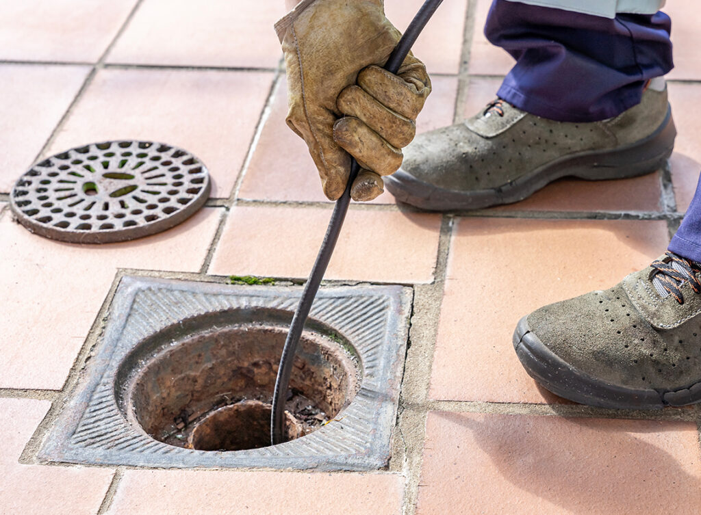 A person wearing gloves and work boots uses a plumbing snake to unclog an outdoor drain on a tiled surface.