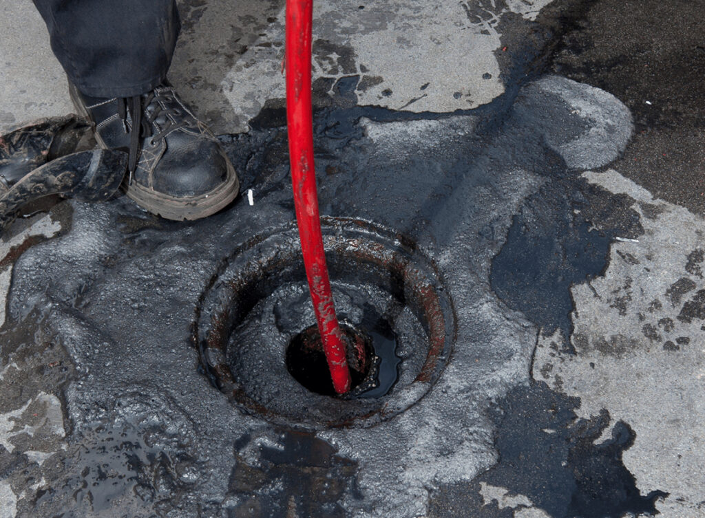 A person wearing black boots uses a red plumbing snake to clear a clogged drain, surrounded by dirty water and oil stains on the concrete surface.