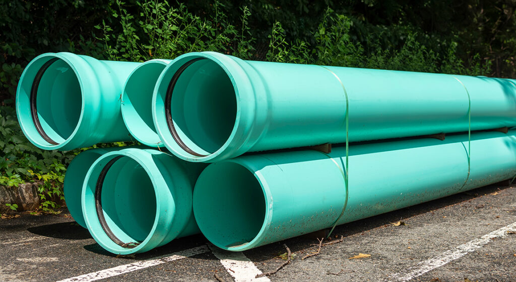 Six large turquoise pipes are stacked in two rows on the ground near a road, surrounded by greenery and trees in the background.