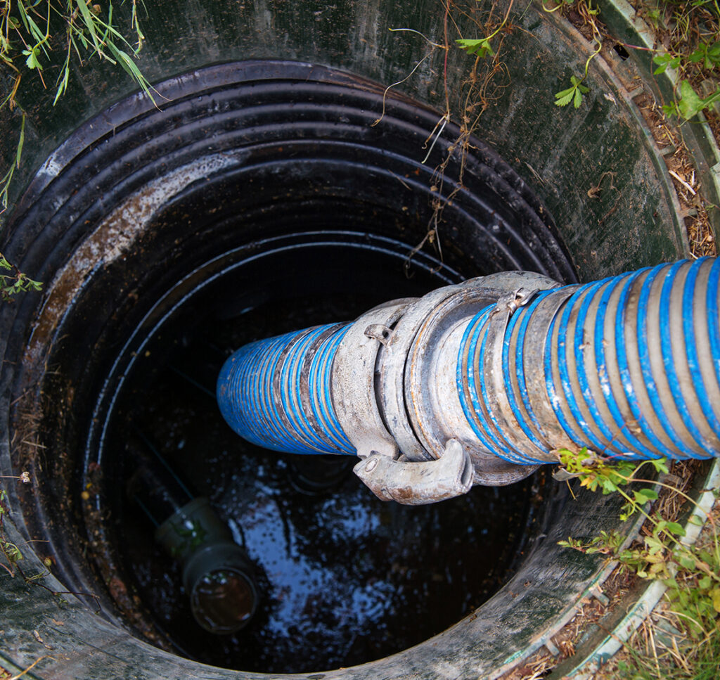 A blue hose extends into an outdoor well or manhole surrounded by grass, with water visible at the bottom and some pipes or equipment partially submerged.