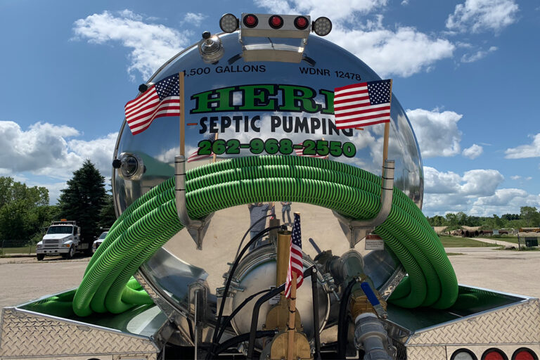 A shiny septic pumping truck with green hoses, American flags, and the text HERR SEPTIC PUMPING 1,500 GALLONS 262-968-2550 on the tank, parked outdoors under a blue sky with clouds.