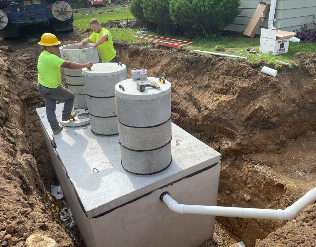 Two workers in safety gear install large concrete septic tank sections into an excavated pit, surrounded by dirt, tools, pipes, and nearby construction materials.
