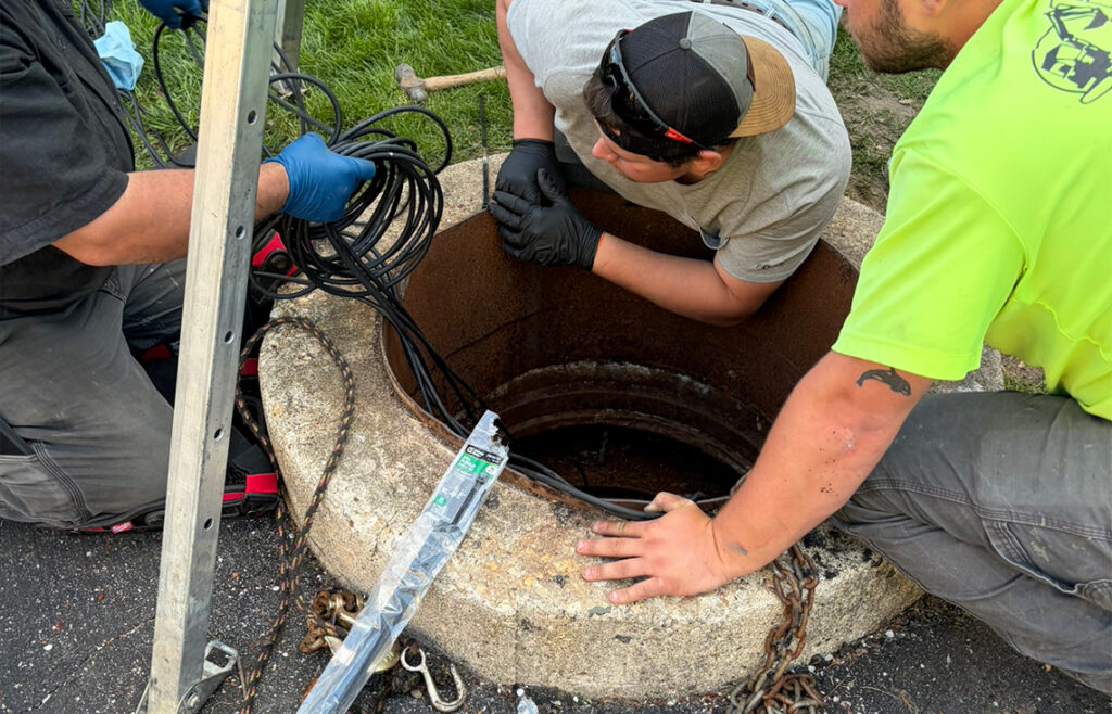Three workers, one holding cables, gather around an open manhole. One worker leans in, inspecting inside, while another steadies cables. Tools and chains are visible around the concrete opening. Grass and asphalt surround the area.