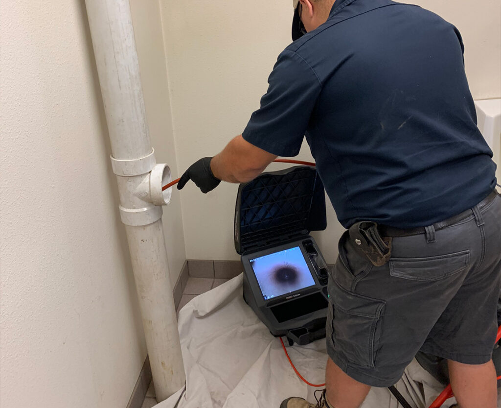 A worker uses a sewer camera inspection tool to look inside a white pipe; the camera feed is visible on a monitor showing the pipes interior.