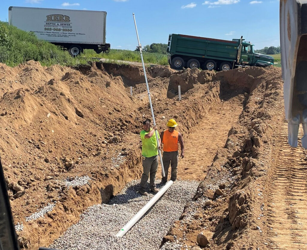 Two construction workers stand in a trench placing a long white pipe on gravel, surrounded by dirt piles. A green dump truck and a white truck are parked on higher ground in the background under a blue sky.