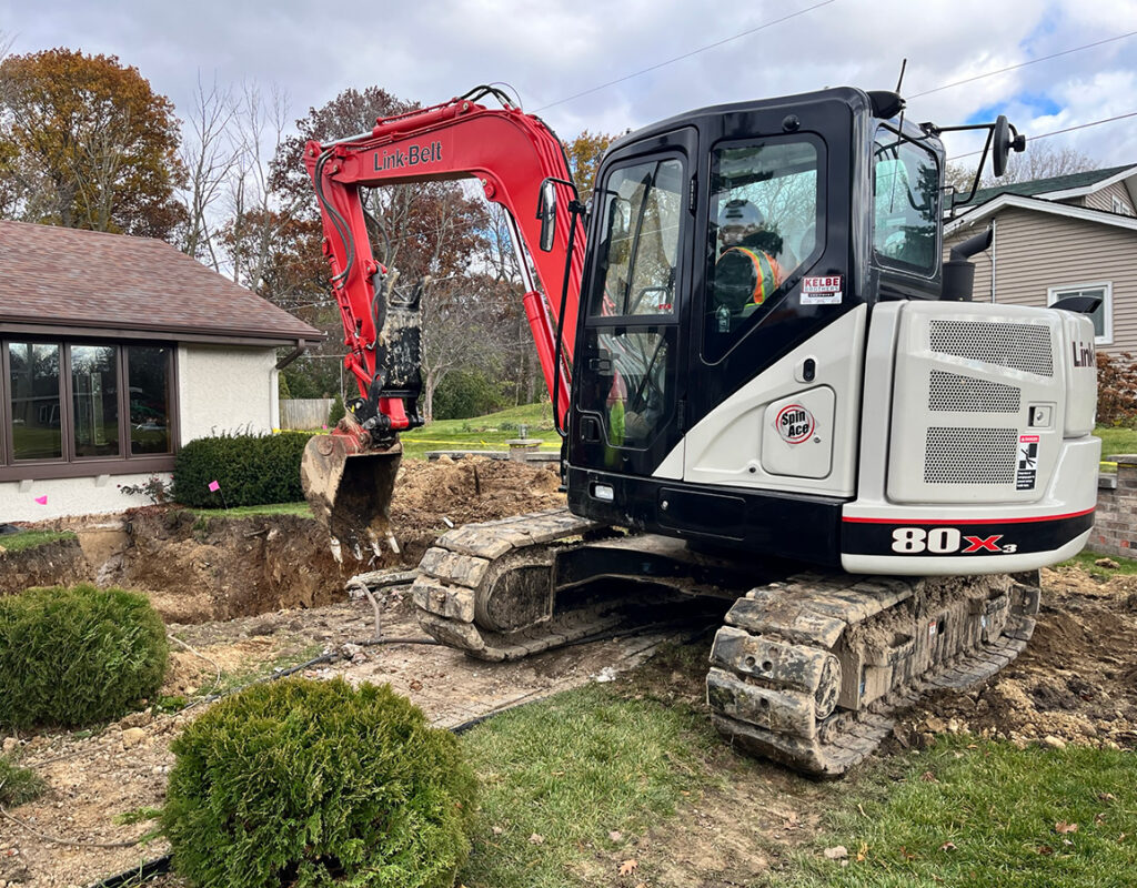A red and white excavator is digging near a house, surrounded by dirt, grass, and bushes. Trees with autumn leaves are in the background under a cloudy sky.