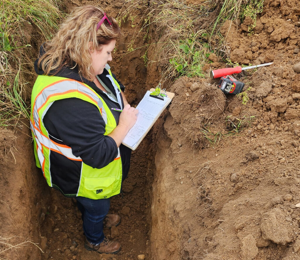 A person in a safety vest stands in a deep soil trench, writing notes on a clipboard. Tools are placed on the soil edge above. The person is surrounded by dirt walls and grass is visible at the top.