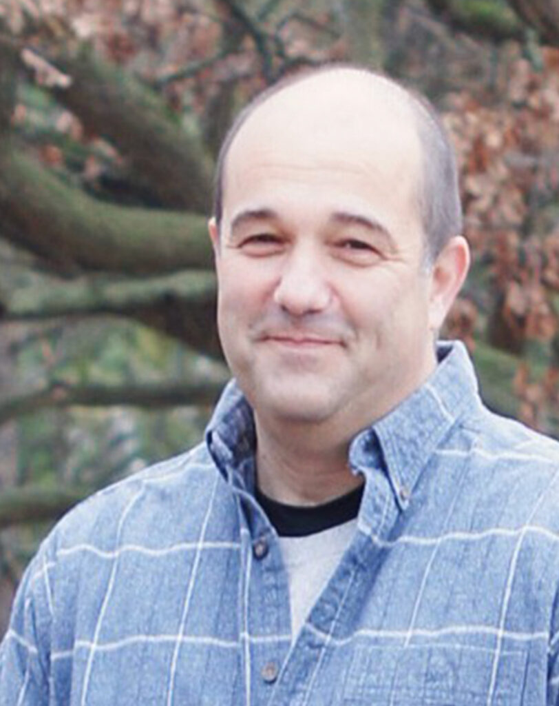 A middle-aged man with short hair and a receding hairline smiles outdoors, wearing a blue plaid shirt over a gray shirt. Trees with brown leaves are visible in the blurred background.