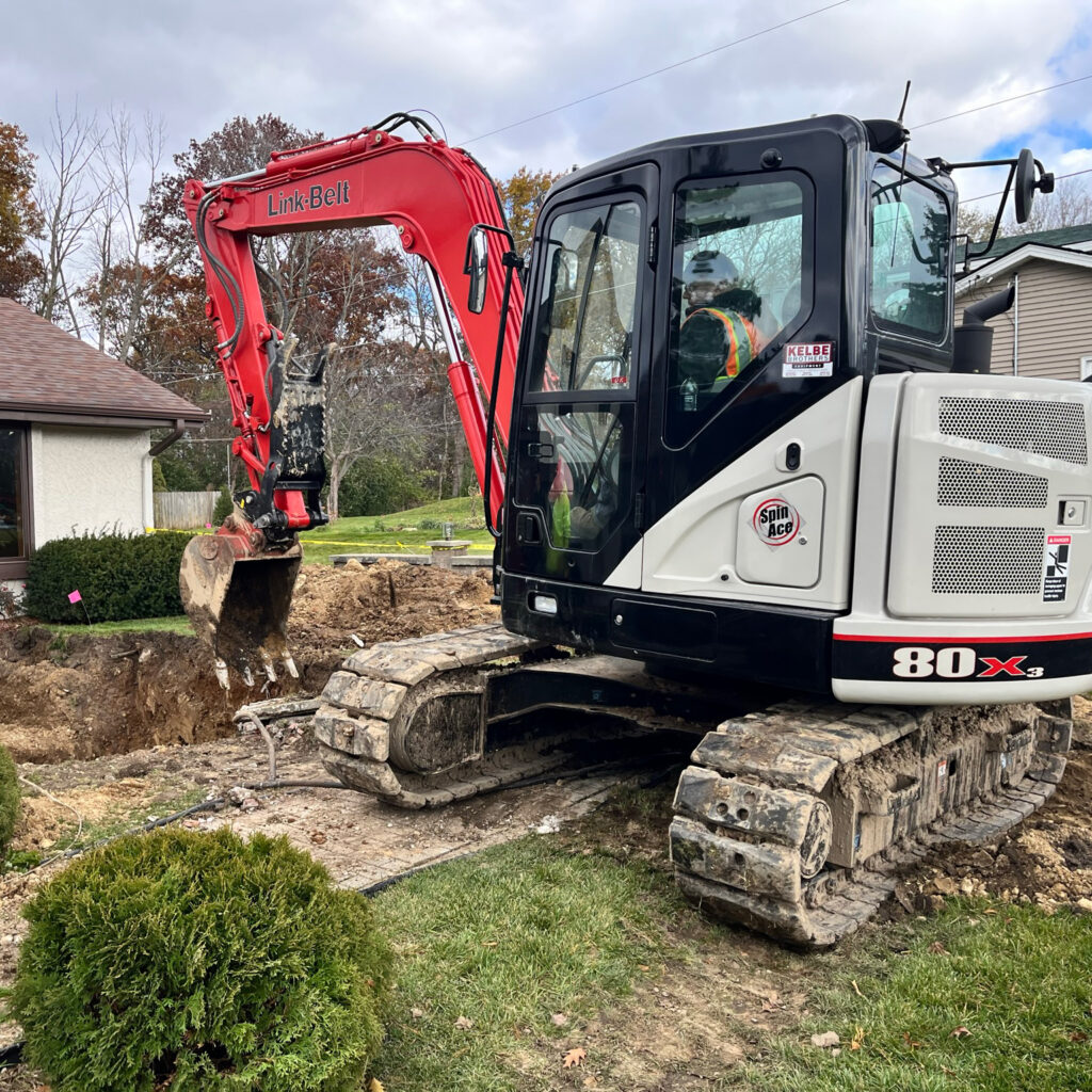 A red and white excavator is digging near a house, with dirt piles and exposed ground visible. The excavator is on tracks, and green bushes and grass surround the work area.